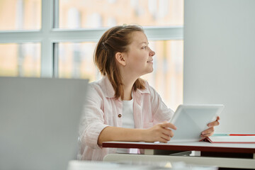 Side view of smiling teen schoolgirl holding digital tablet and looking away during lesson in class
