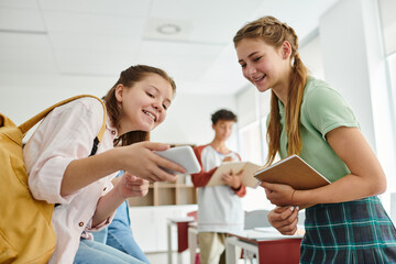 Smiling teen schoolgirls with backpack and notebook using smartphone in classroom in school