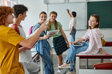 Smiling schoolboys talking and pointing with finger near schoolgirls with devices in classroom