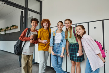 cheerful teenage students holding devices and looking at camera in school hallway, teen friends