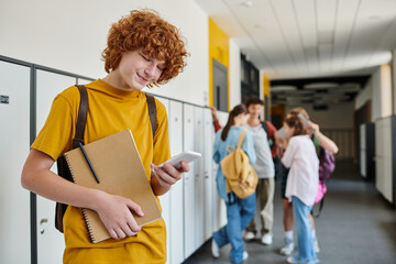 happy redhead schoolboy using smartphone and holding notebook, blur, students in school hallway