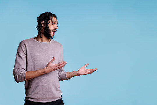 Arab man pointing with hands to side while talking and advertising product. Young adult arabian person with braided black hair showing to left with arms on blue background