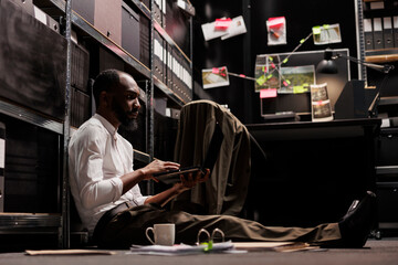 African american cop sitting on floor and working on laptop in office at night time. Police investigator examining clues and suspect photos on computer to solve crime case