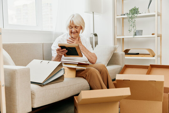 elderly woman sits on a sofa at home with boxes. collecting things with memories and moving and happiness smile.