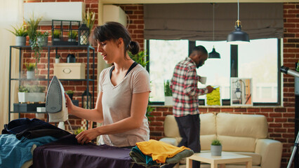Cheerful woman dancing and having fun ironing clothes in living room, girlfriend listening to music and singing at home. Happy adult doing housework and using iron with steam after laundry.
