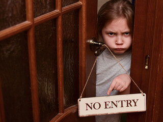 An angry child closes the door to his room and points to a sign that says No entry. The child does not let her parents into the room. Upbringing problems, the conquest of personal space by children