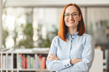 Portrait of beautiful smiling confident businesswoman, manager, CEO holding arms crossed looking at camera. Happy smart university student wearing eyeglasses standing in modern library, education - obrazy, fototapety, plakaty