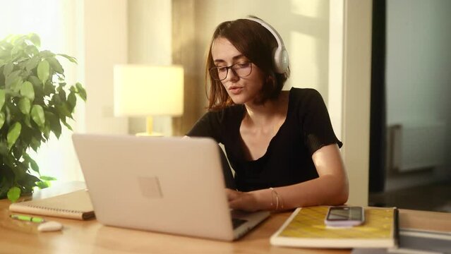 Pretty young woman freelancer with headphones listening music while working online typing browsing on laptop and singing indoors Positive female enjoying great working day at cozy home