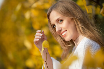 Beautiful young woman with light brown hair in a white sweater on a background of foliage in an...