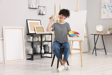 Young woman with brush and artist`s palette near easel in studio