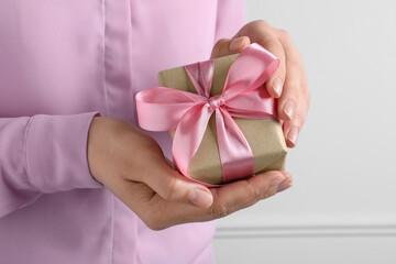 Woman holding gift box with pink bow near white wall, closeup