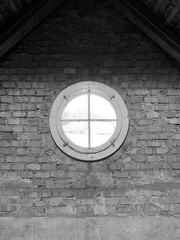 Schwarz-weiß-Aufnahme eines runden Fensters auf einem Dachboden mit Backsteinwand