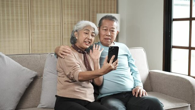 Middle-aged Aaian couple using smartphone together or making video call while sitting at sofa in cozy living room.