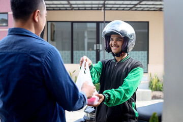 Online food delivery with motorbike wearing green jacket and helmet, holding a bag of food delivering it to customer.