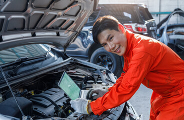 Fototapeta na wymiar Young Asian mechanic in orange uniform View on laptop about car engine while repairing in garage