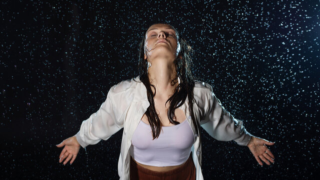 young woman in the rain, getting wet through, wet clothes and water running down her face. a beautiful brunette stands with her face exposed to the streams of water, portrait in aqua studio