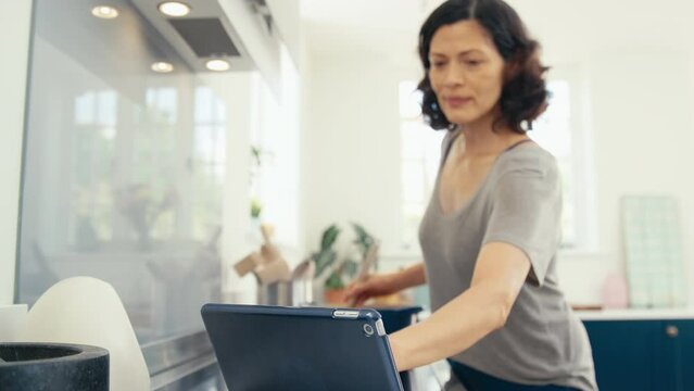 Mature woman cooking meal in kitchen at home following online recipe on digital tablet - shot in slow motion