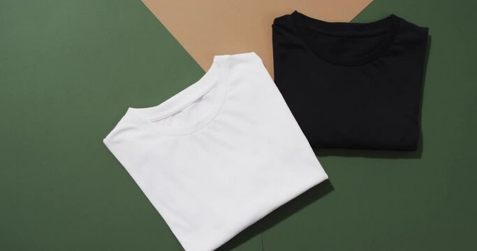 Video of flat lay of folded white and black t shirts with copy space on green and brown background