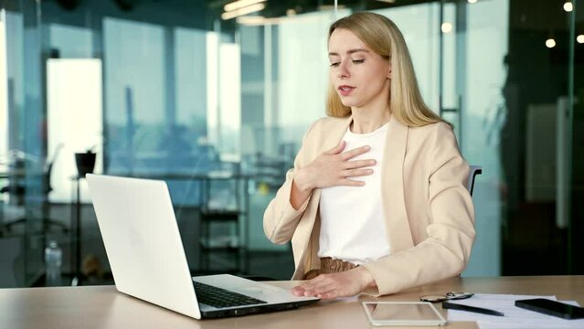 A young businesswoman having a panic attack while working on a laptop while sitting at a workplace at a desk in a modern office. The employee chokes, holds his hands to his chest, breathes hard