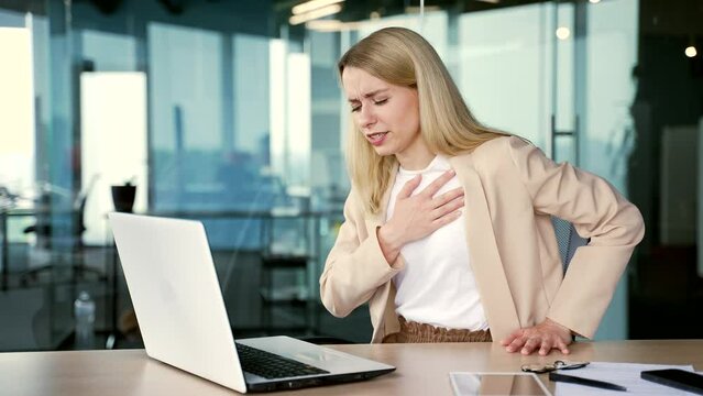 Sick young businesswoman suffering from chest tension, heart attack while working on laptop while sitting at desk at workplace in modern office. A woman in a suit needs urgent medical attention