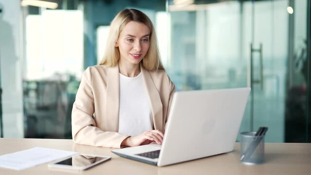 A young businesswoman typing on a laptop while sitting at a desk at a workplace in a modern office. Blonde female employee working on a project on the computer, texting a client or chatting online