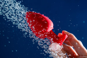 Woman washing red anal plug under shower on blue background. 
