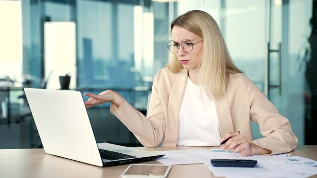 Confused young female accountant having difficulty with work on a laptop while sitting in office. A sad businesswoman looks at the computer screen, spreads her hands and cannot understand the problem