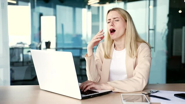 Overworked young female employee feels tired and wants to sleep working on laptop while sitting at workplace at desk in modern office. Sleepy exhausted woman yawns, closes her eyes. Overload at work