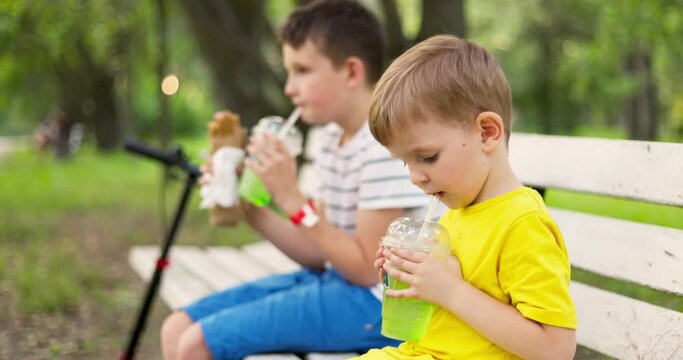 Children drink soda beverage and eat hot dog sitting on a bench in the park