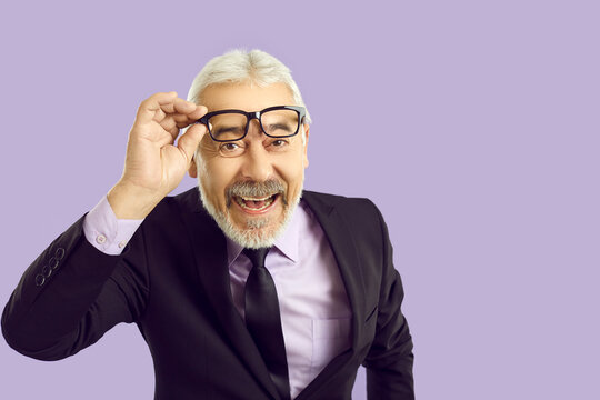 Studio Portrait Of Happy Senior Businessman. Cheerful White Haired Bearded Mature Man In Stylish Suit Lifts His Glasses, Looks At Camera, Smiles And Laughs Standing Isolated On Solid Lilac Background