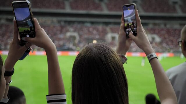 A view from the back of women in the stands of a football stadium, they take photos on their phone. Football fans in the stands of the stadium during a football match.