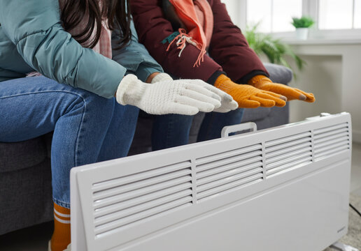 Young Couple Freezing In House With Broken Heating During Cold Winter. Two People Wearing Winter Coats And Gloves Sitting On Couch At Home And Warming Up Hands By Electric Heater
