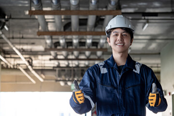 Portrait of Asian confident man construction worker in protective uniform suit and safety helmet showing thumbs up in unfinished building. Male engineer showing hand gesture sign at construction site