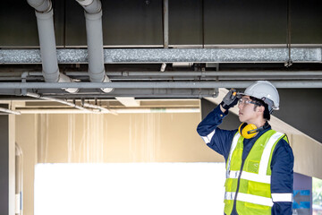 Confident man construction worker with reflective vest, protective goggles and ear muffs. Asian male maintenance engineer holding his safety helmet while looking at piping system at construction site