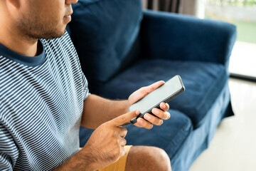 Close up male hands using smartphone sitting on blue sofa in the house. Man with typing cellphone sending message while sitting on couch in living room