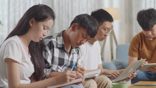 Close Up Of Asian Teen Group Studying At Home. Helping Each Other Doing Project, A Boy In Plaid Shirt Thinking, Raising Index Finger, Sharing Idea With Friends, Writing Into Notebook And Typing On A L