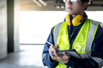 Building inspector man using digital tablet checking safety and security system. Asian male worker in reflective vest and protective ear muffs working for building maintenance inspection
