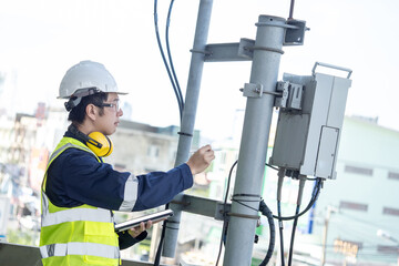 Building inspector man using digital tablet while checking telecommunication pole or telecom tower. Asian male technician inspecting and pointing at cellular phone network antenna on building rooftop - obrazy, fototapety, plakaty