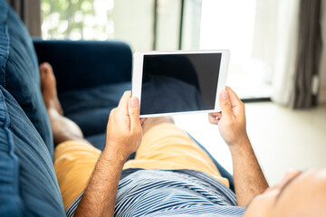 Close up male hands using tablet lay down on blue sofa in the house. Man with typing cell phone sending message while lying down couch in living room