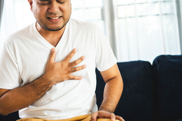 Young Asian man he felt a pain in his chest held his hand and squeezed it in living room. Heart...