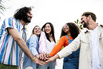 Diverse group of cheerful millennial friends standing together stacking hands in the street. Cheerful multiethnic young carefree friends laughing and having fun outside joining hands in the center.