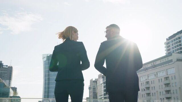 Back view following two positive partners businessman businesswoman talking and smiles walking outdoors along terrace modern office building roof overlooking cityscape lit with morning sunshine light