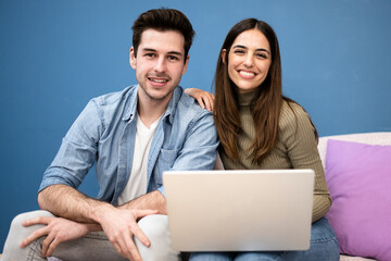 happy young couple sit on couch and using laptop and looking at camera. Cheerful couple smiling and searching on computer