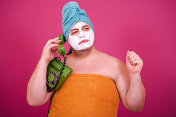 Early morning. Funny fat man talking on a retro phone. The guy wrapped in a towel after a shower poses on a pink background.