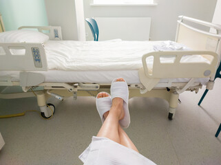 Female legs in white slippers in a hospital ward against the background of an empty hospital bed.