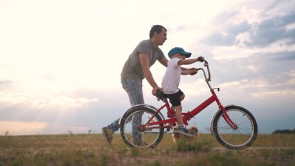 Fototapeta na wymiar dad teaches son to ride a bike. happy family kid dream concept. the boy sat on bicycle for the first time, his father teaches boy to ride a bicycle. dog runs with family, lifestyle fun family pastime