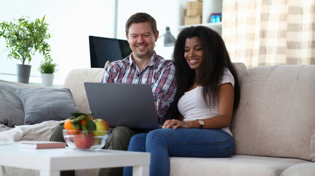 Portrait of happy smiling couple viewing photos, watching movie on laptop. Man and african-american woman sitting on cozy sofa. Home interior and mixed race marriage concept