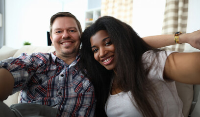 Portrait of cheerful mixed race couple taking happy selfie on sofa indoors. Man and latino woman widely smiling on camera. Multinational and biracial family concept