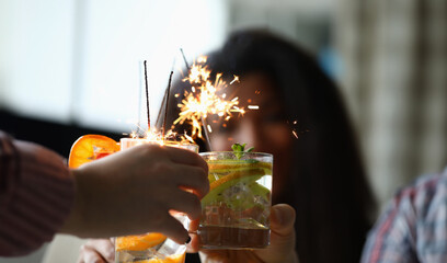 Close-up of friends hands holding fruit cocktails with sparklers decoration. Delicious refreshing beverage. Christmas, new year celebration party and holiday concept