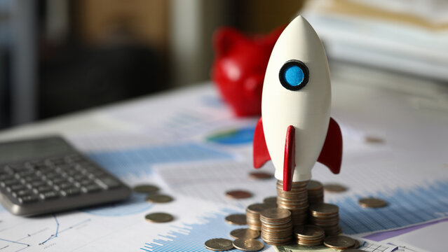 Close-up of rocket on pile of golden coins symbolizing company profits growth. Money scattered all around important charts and graphs. Start up and development concept